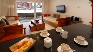 Habitación de hotel con sofá, cama, televisor y zona de comedor con cruasanes y café.