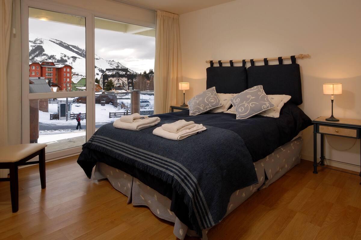 Habitación de hotel con una cama grande, dos mesitas de noche, una silla y vista a un paisaje nevado a través de un ventanal amplio.