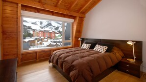 Un dormitorio de cabaña de madera con un ventanal grande, una cama con ropa de cama marrón, una mesita de noche y una lámpara.