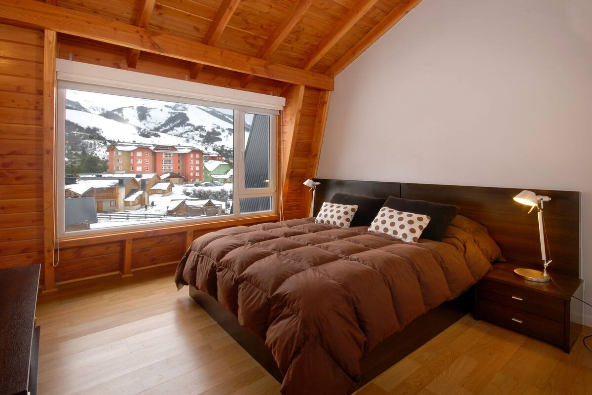 Un dormitorio de cabaña de madera con un ventanal grande, una cama con ropa de cama marrón, una mesita de noche y una lámpara.