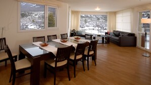 Un comedor con mesa de vidrio y sillas de madera, un sofá, y vista a un paisaje nevado a través de grandes ventanales.