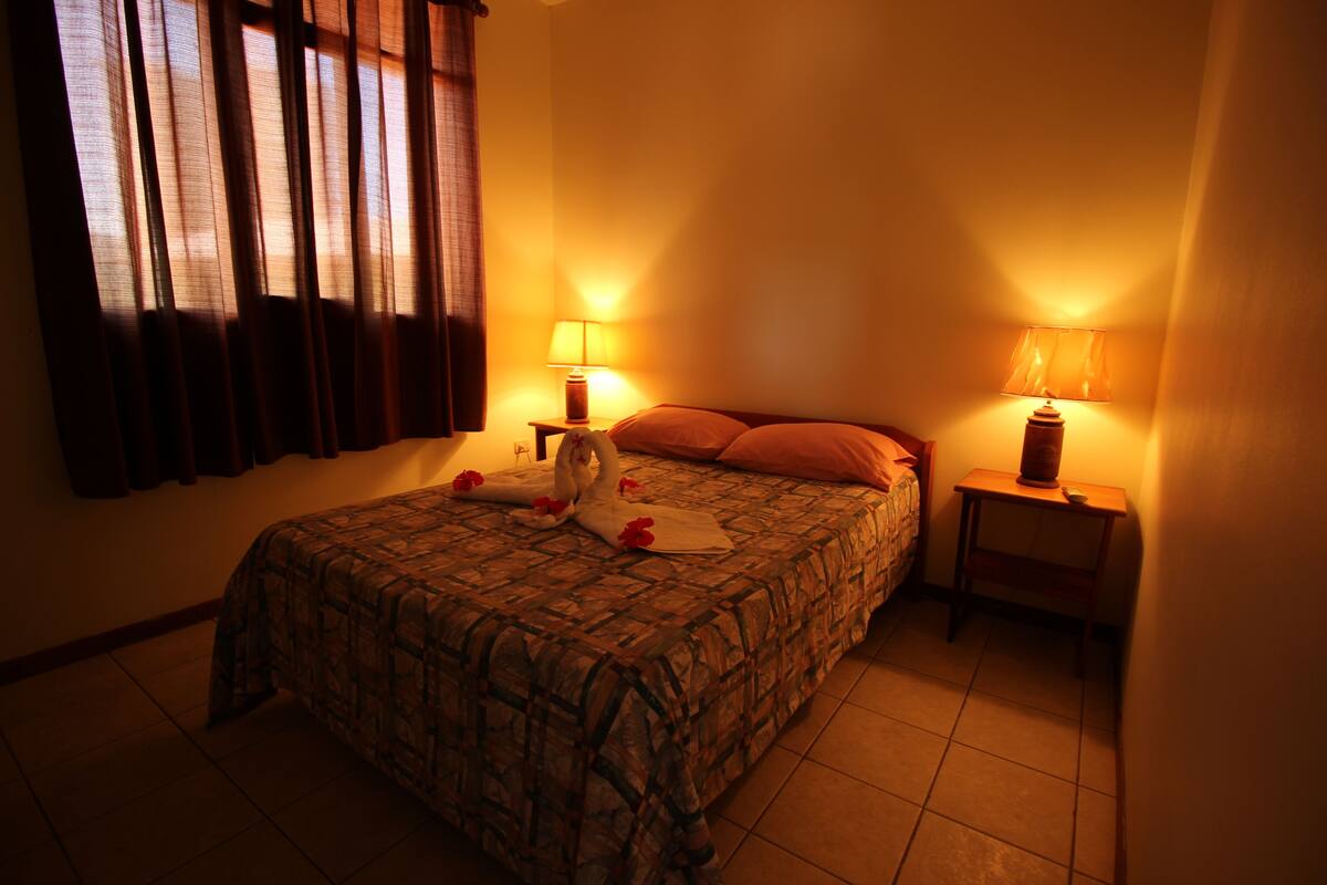 A bedroom with a bed, two bedside lamps, a window with curtains, and a bedside table.