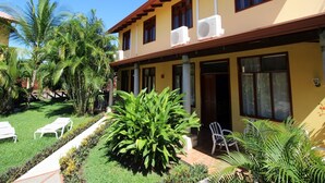 Um edifício de dois andares com fachada amarela, grandes janelas e unidades de ar condicionado no telhado. Possui um jardim bem cuidado com palmeiras e um caminho que leva à entrada.