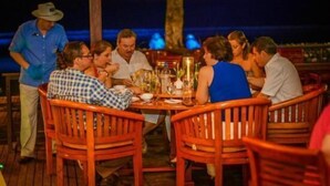Um grupo de pessoas jantando em um restaurante ao ar livre, com cadeiras e mesas de madeira, com um céu escuro e um corpo d'água ao fundo.