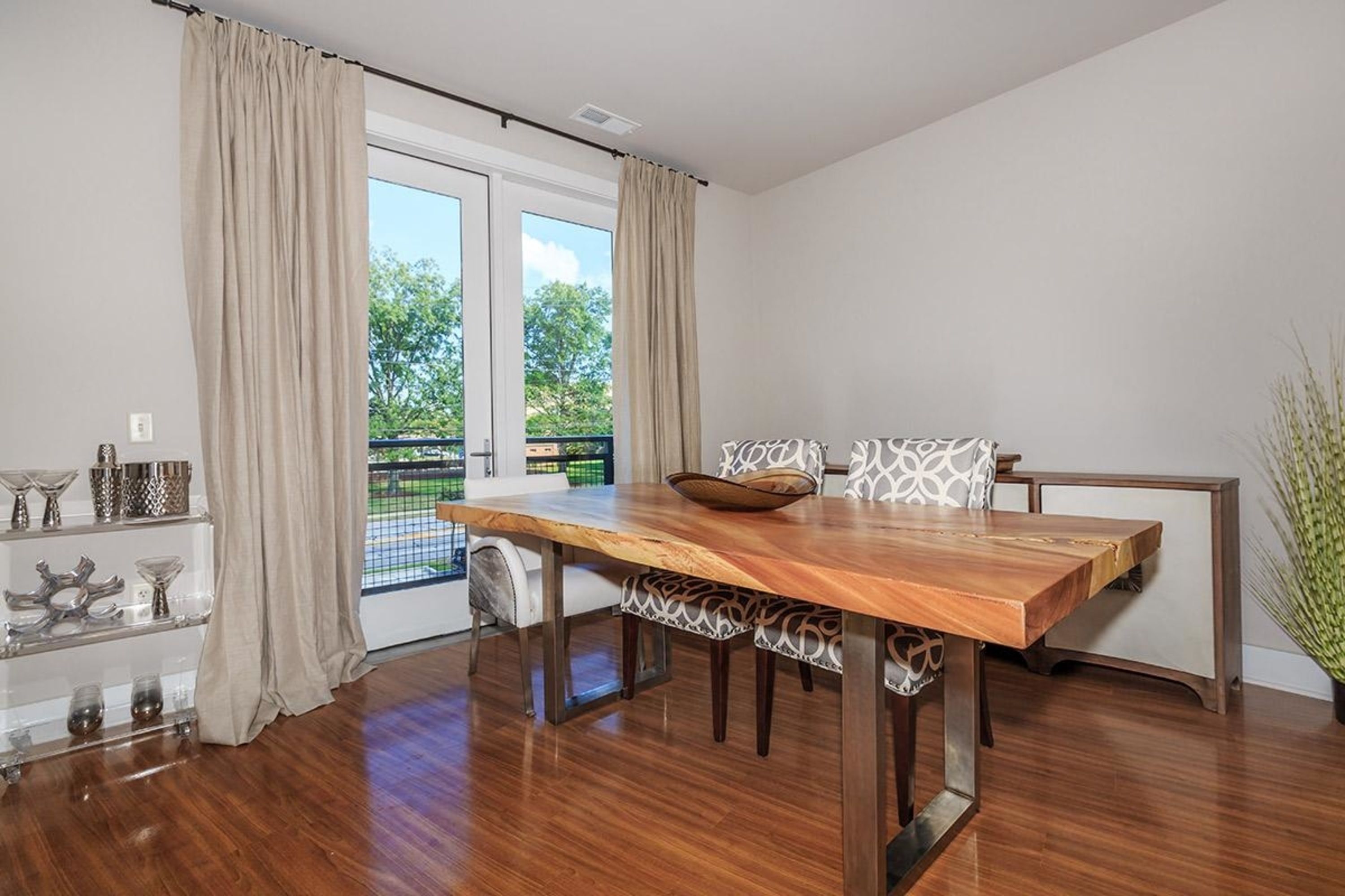 A dining area with a wooden table, white patterned cushions, and a view of greenery through a sliding door.