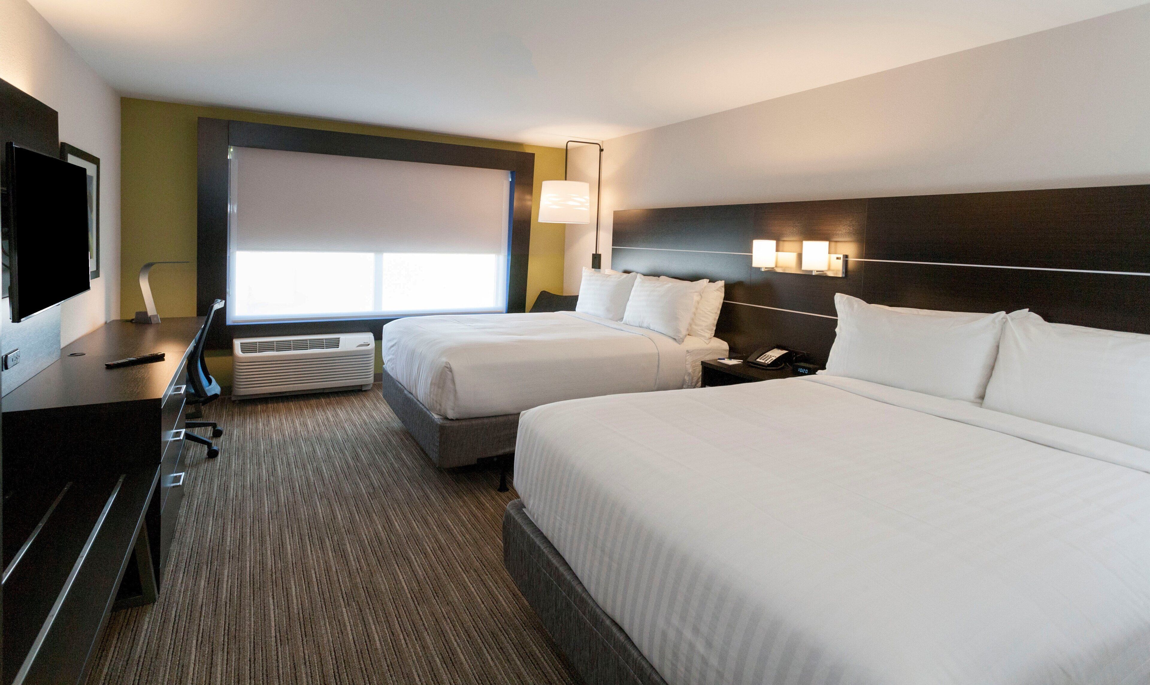 Une chambre d’hôtel avec deux lits, un bureau avec un ordinateur, une télévision et une fenêtre avec des stores.