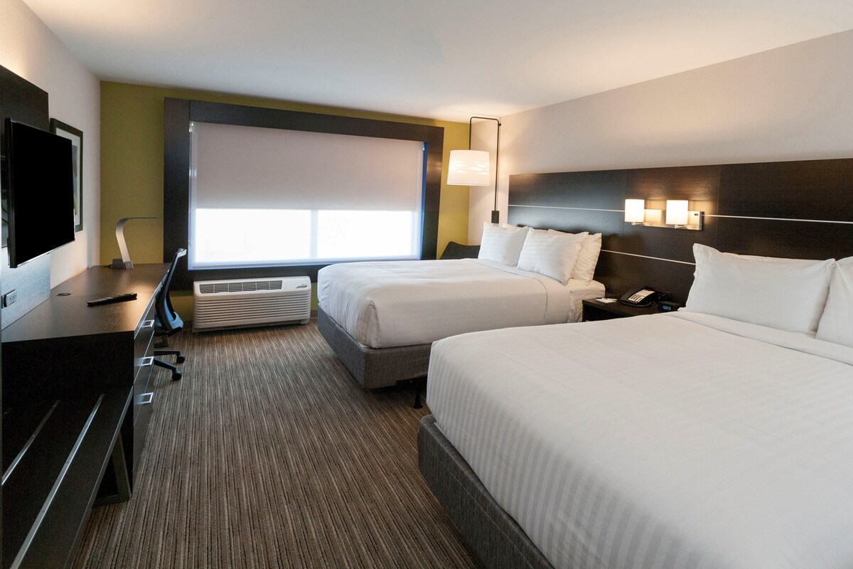 Une chambre d’hôtel avec deux lits, un bureau avec un ordinateur, une télévision et une fenêtre avec des stores.