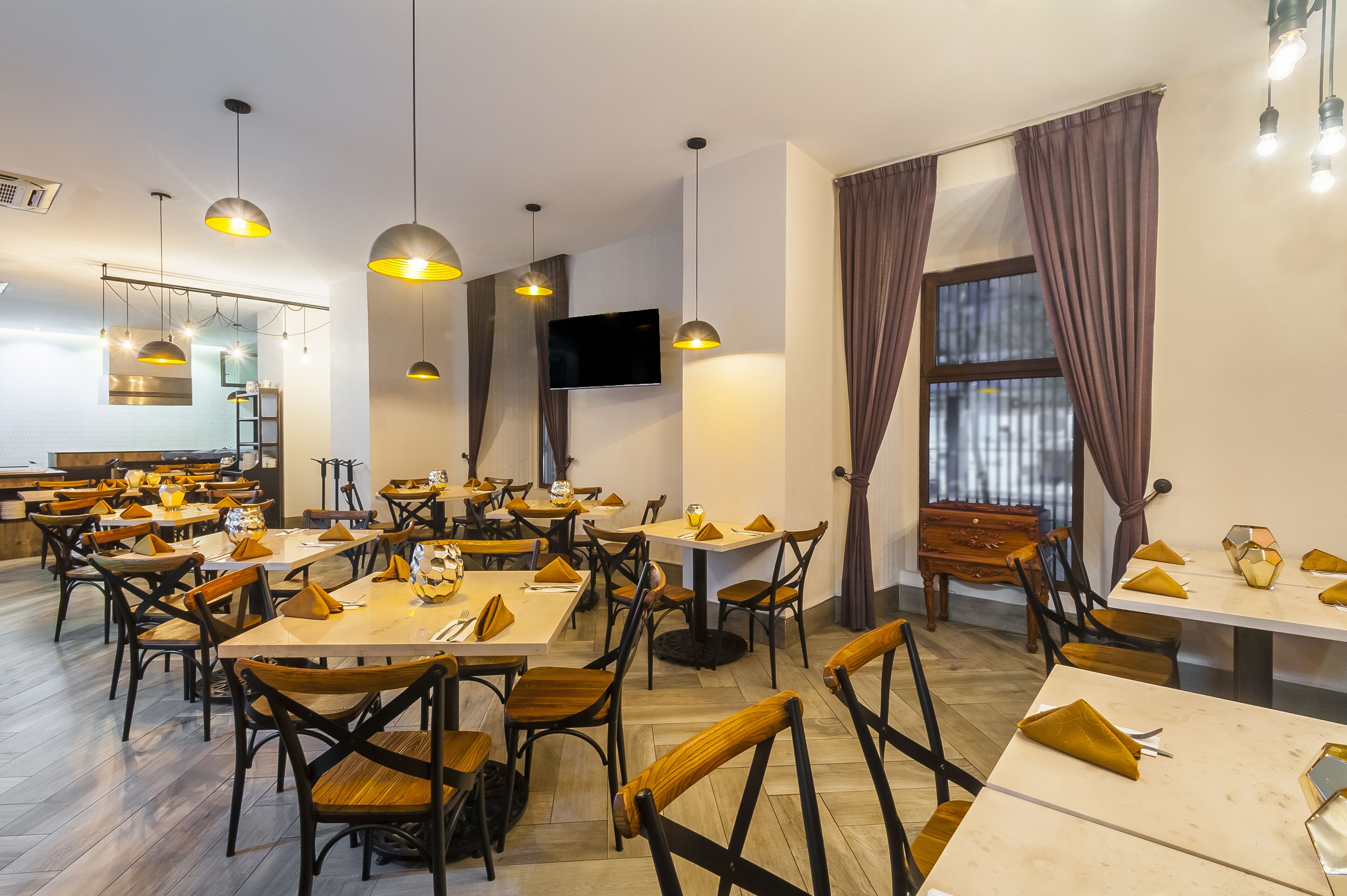 A restaurant with multiple tables and chairs, pendant lighting, a flat-screen TV, and large windows with curtains.