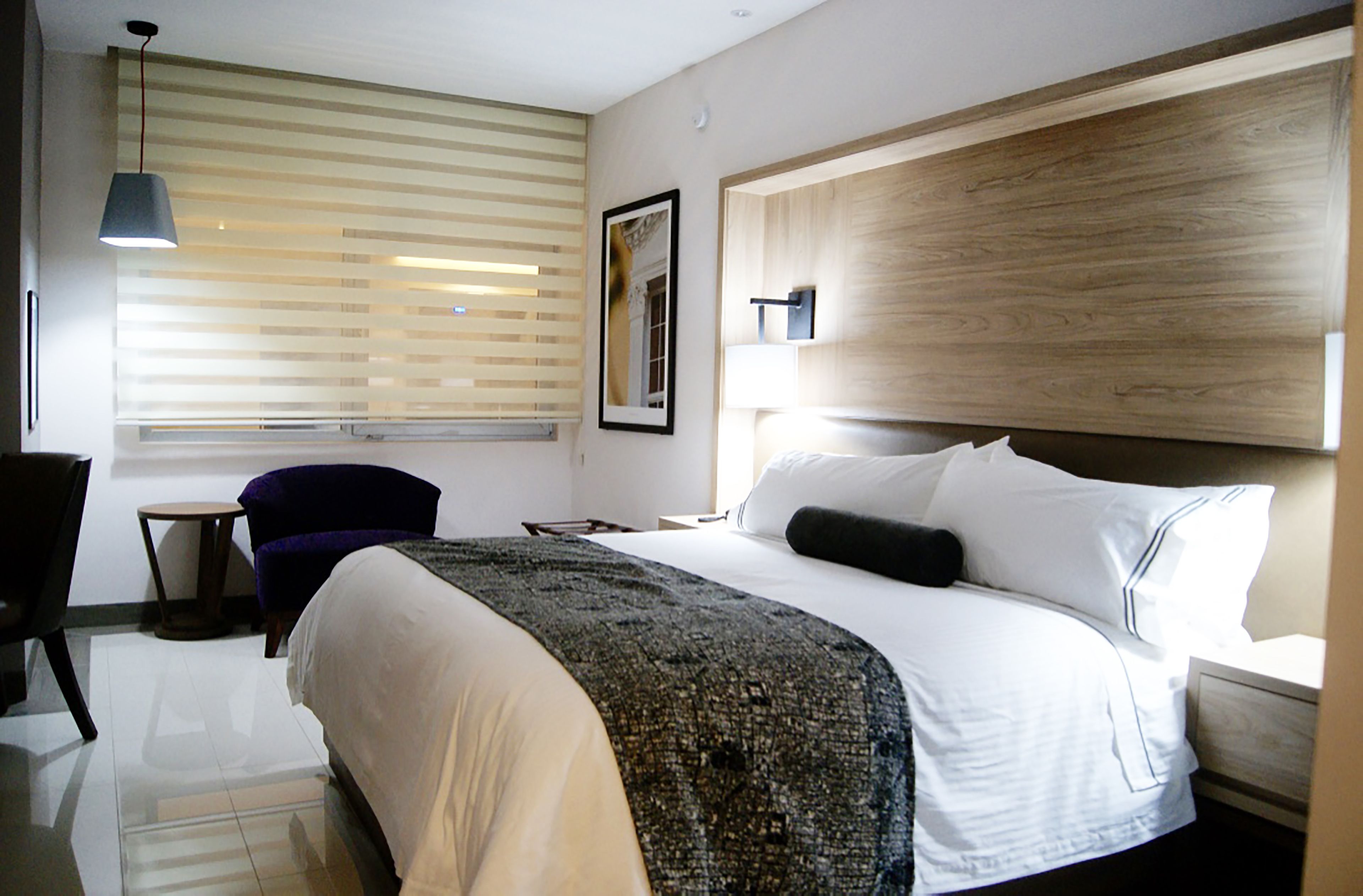 A hotel room with a large bed, a bedside table, a chair, a small table, and a window with blinds.