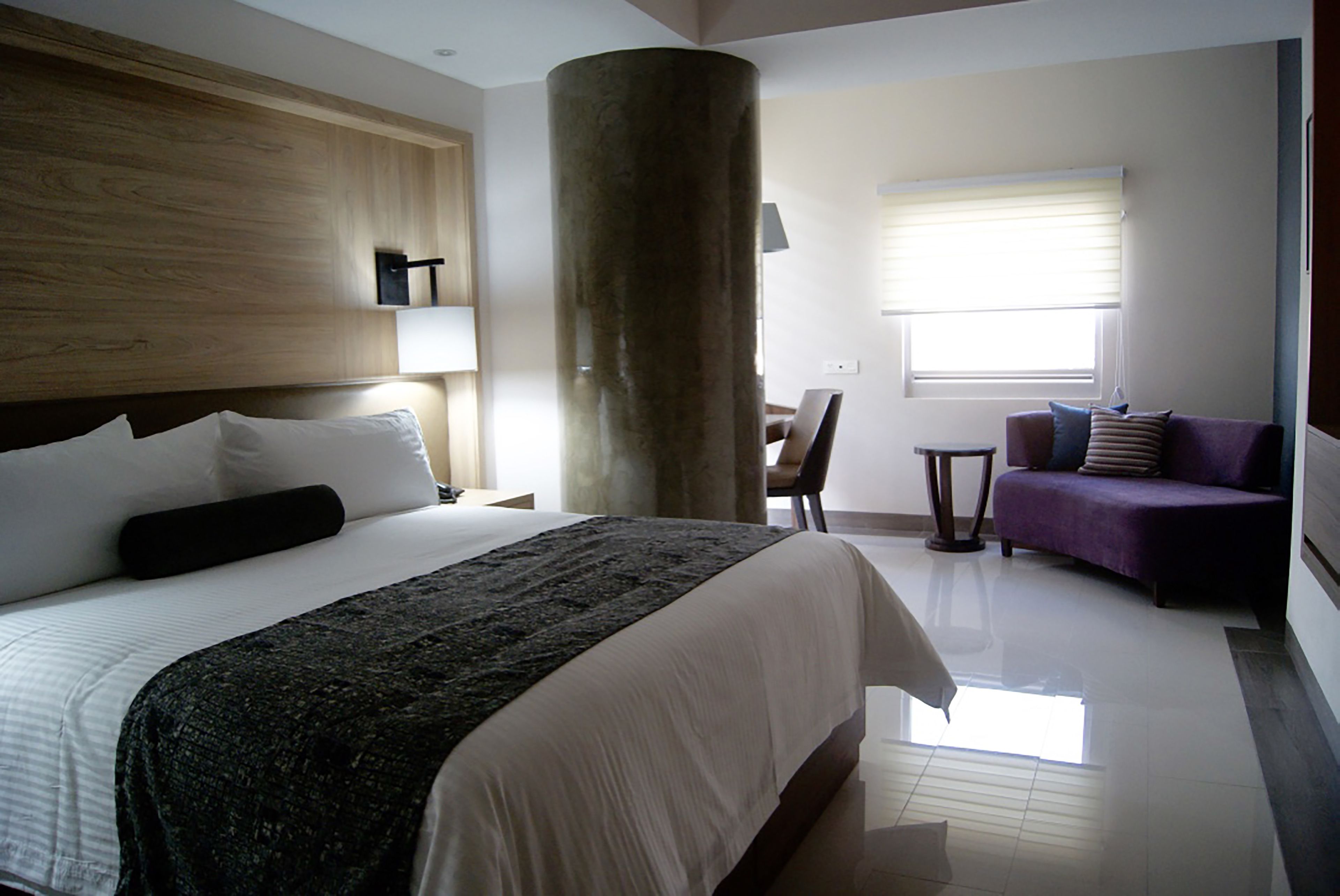 A hotel room with a large bed, a wooden headboard, a large pillar, a purple sofa, and a window with blinds.