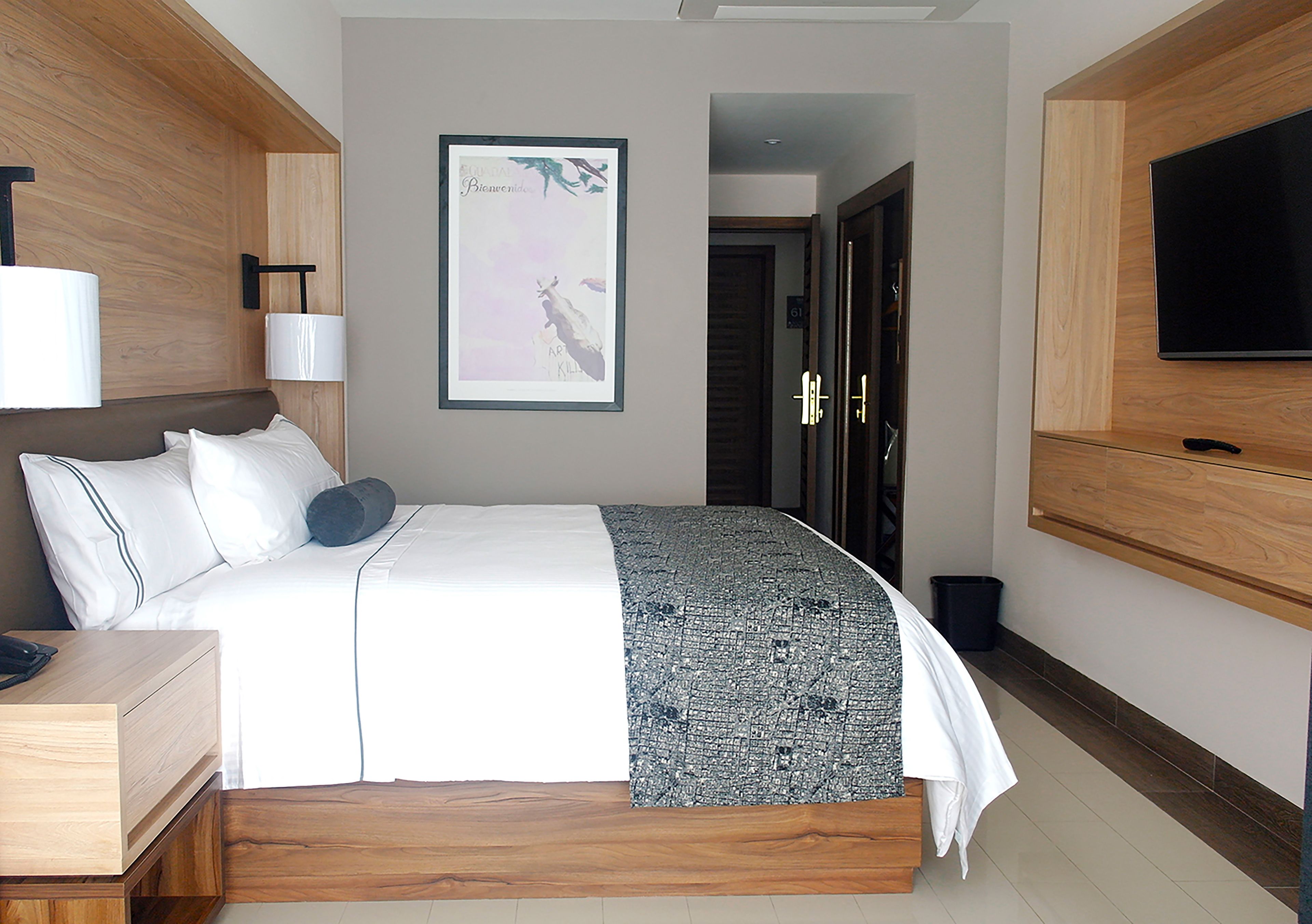 Et moderne hotelværelse med en stor seng, et sengebord, et fjernsyn ophængt på væggen og et indrammet billede over sengen.