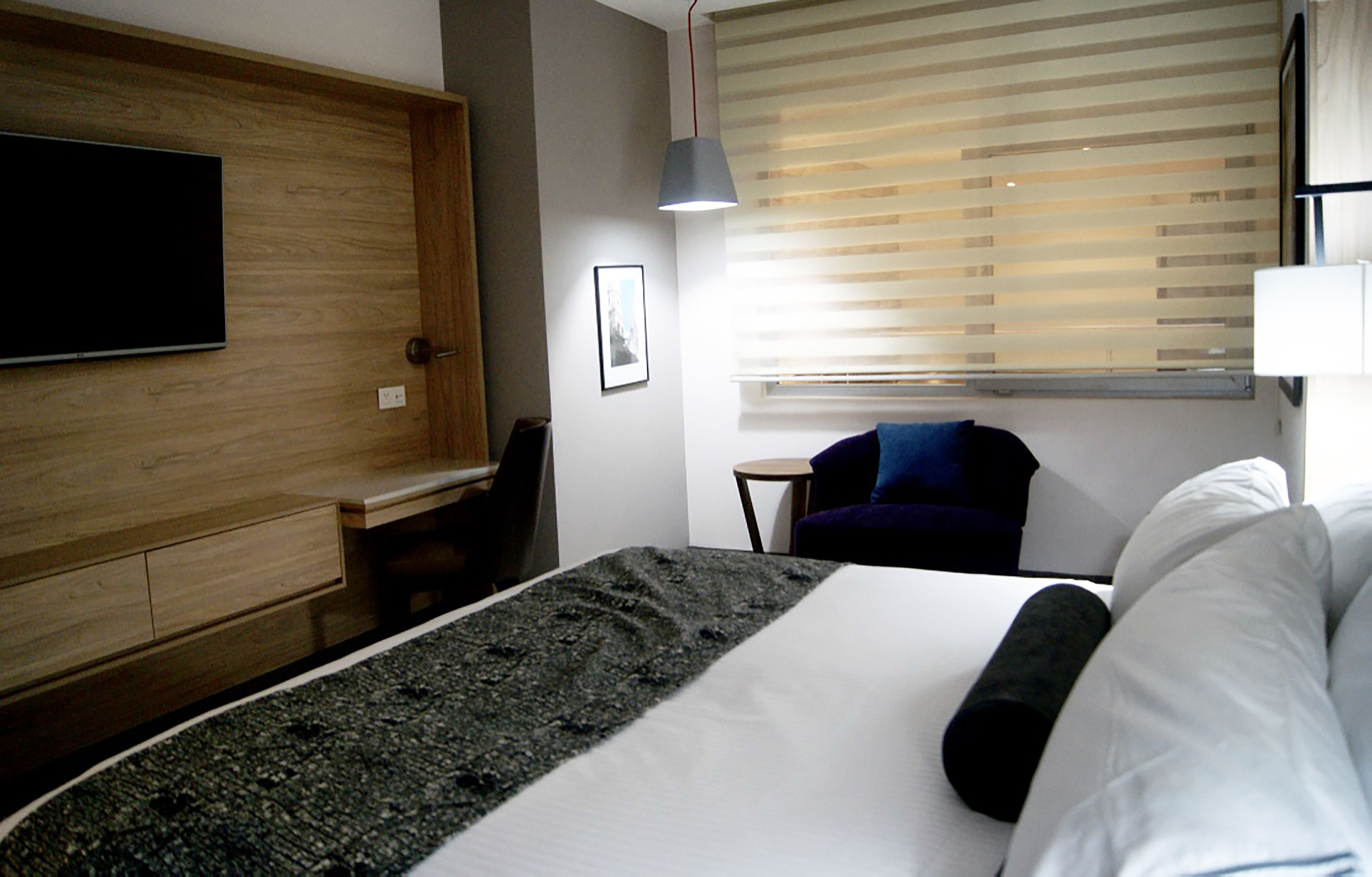 A hotel room with a bed, a desk, a chair, a TV mounted on the wall, and a window with blinds.