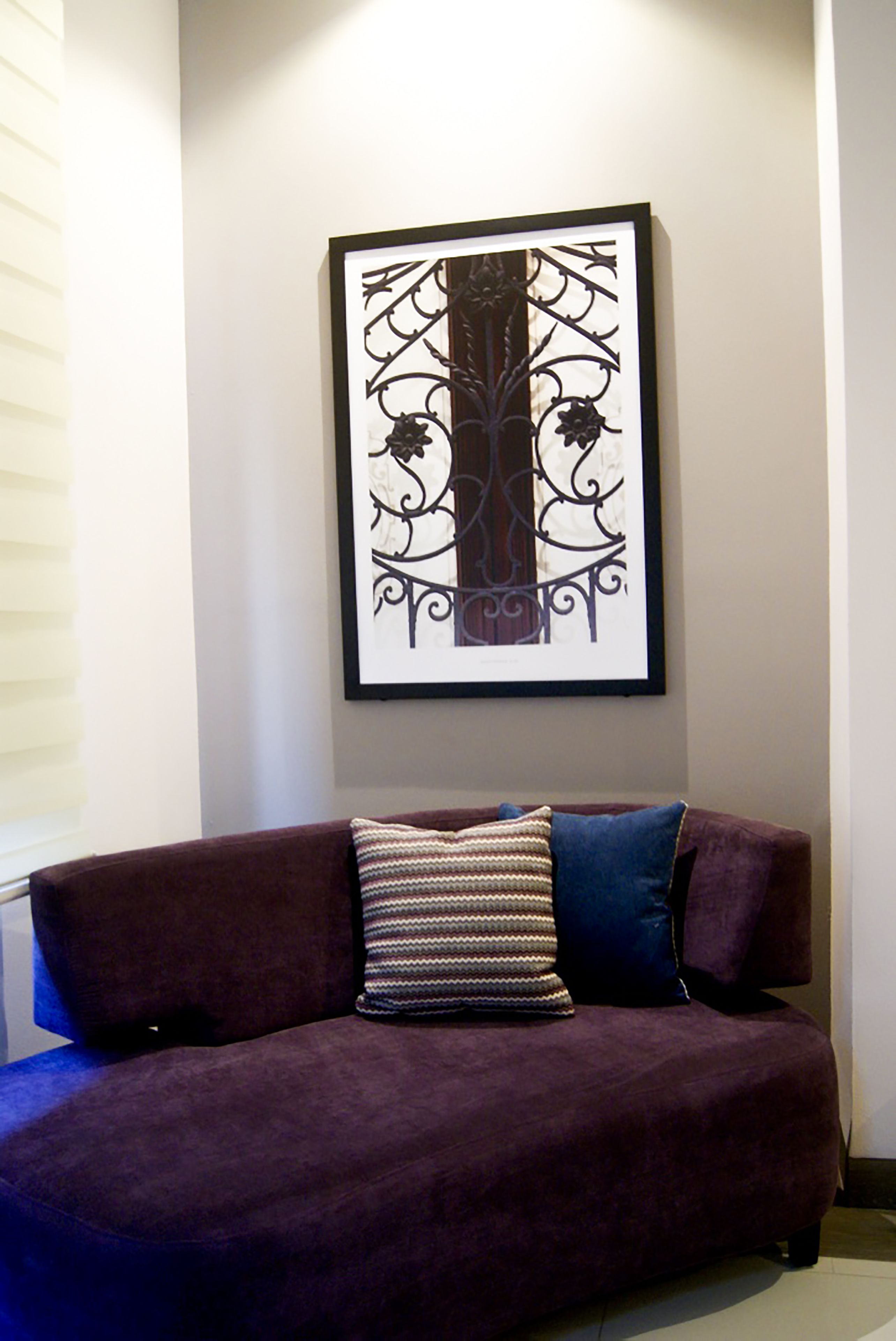 A purple sofa with patterned cushions, a framed artwork above it, and a window with blinds to the left.