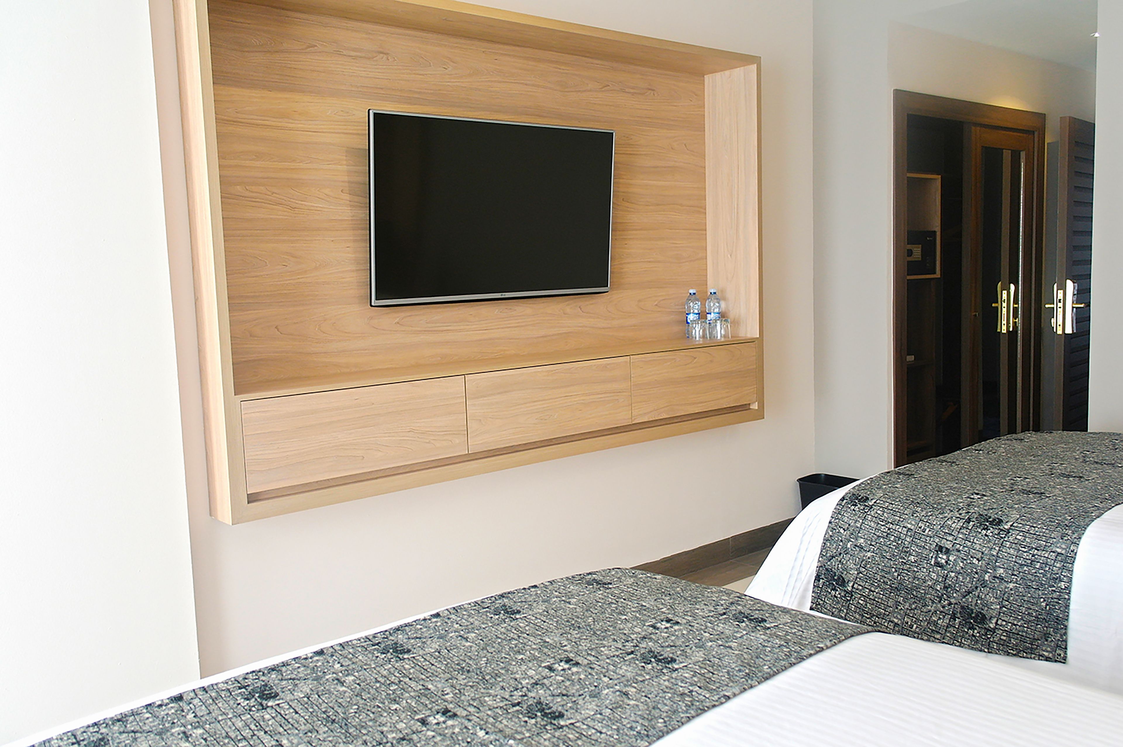 Habitación de hotel con una televisión de pantalla plana montada en un panel de madera, dos mesitas de noche con botellas de agua y una cama con colcha estampada.