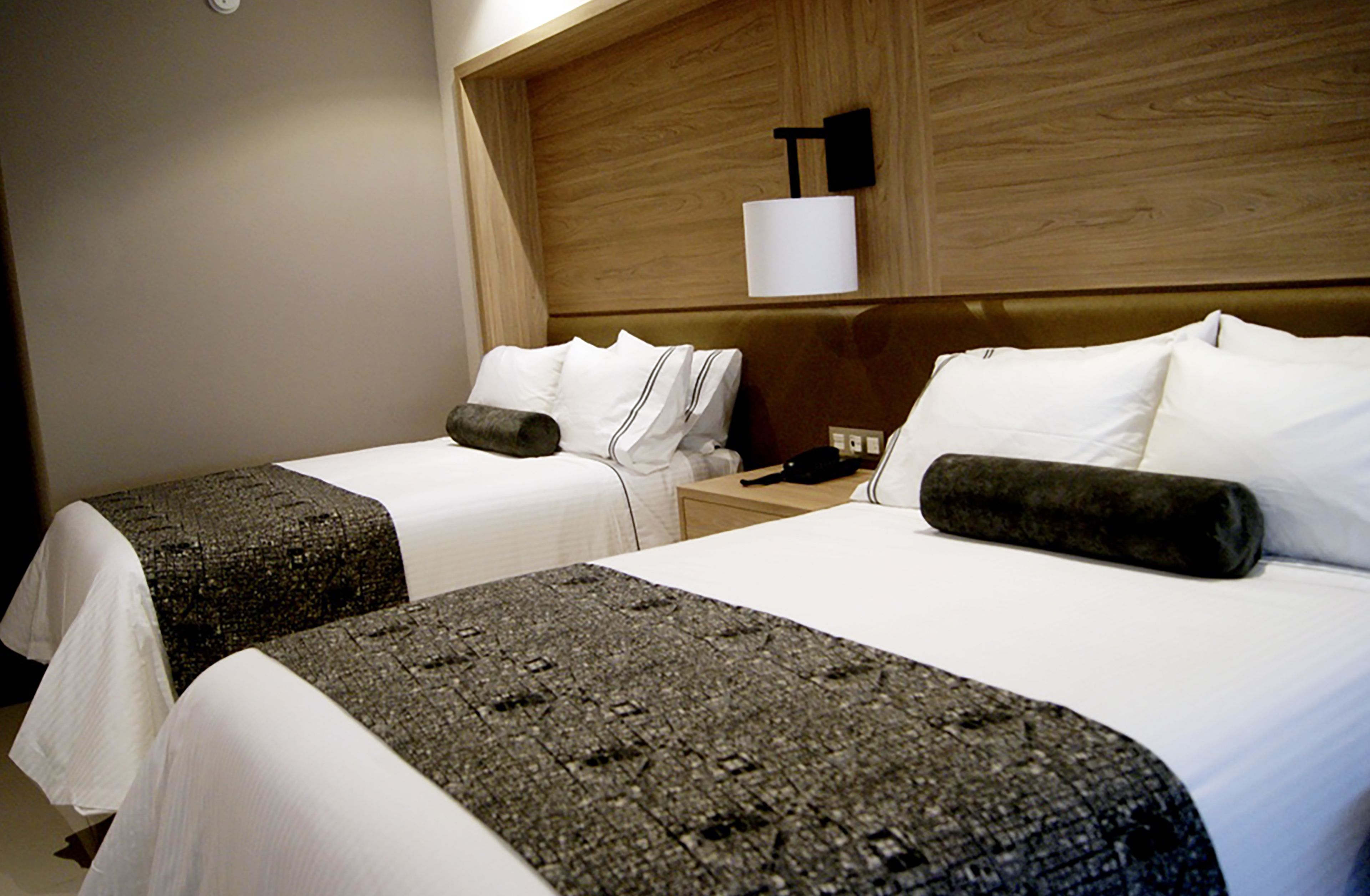 A hotel room with two beds, each with white linens and a dark patterned bedspread. There is a wooden headboard with a wall-mounted lamp and a small table beside each bed.