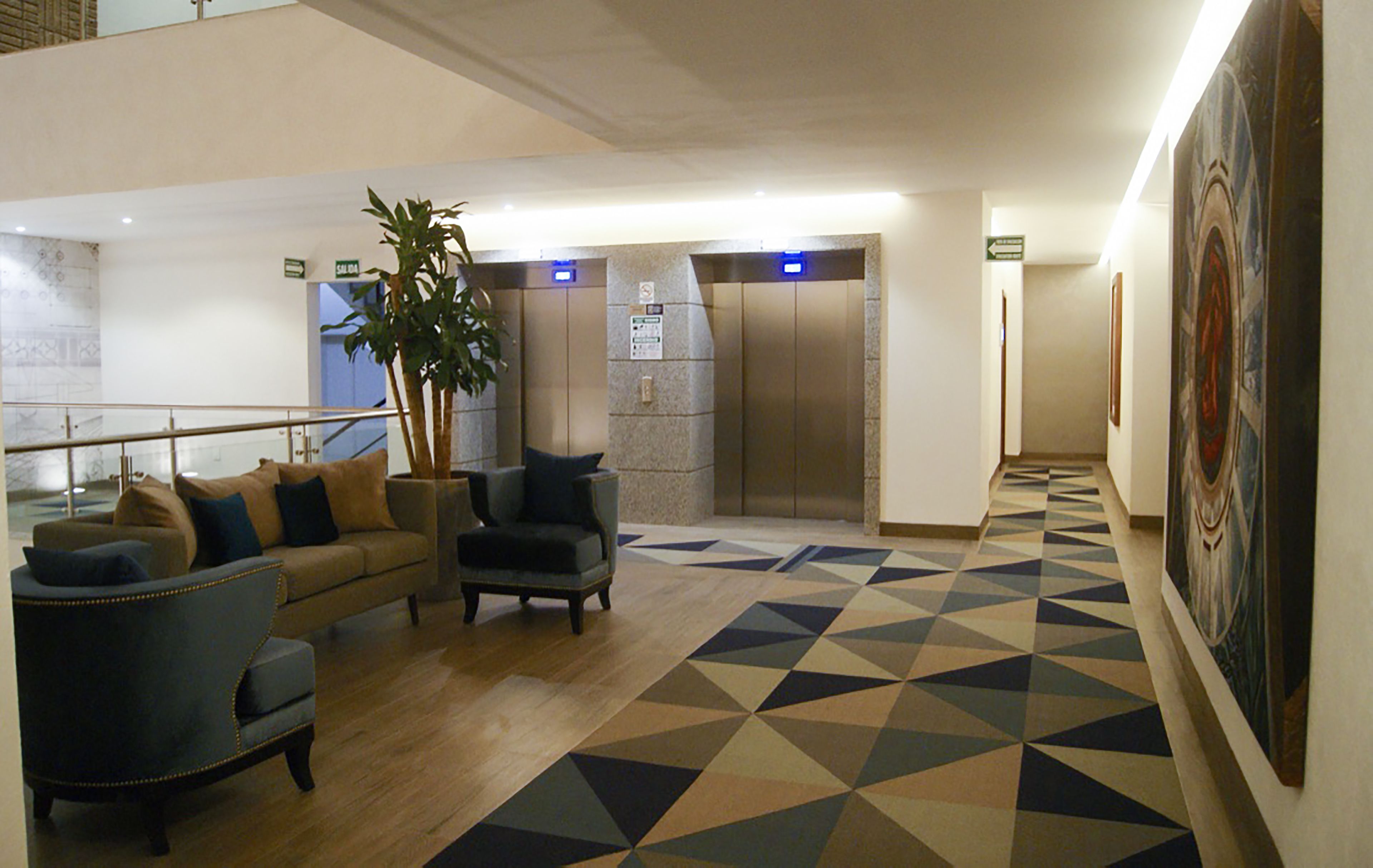 A lobby with a geometric patterned floor, a sofa, armchairs, a potted plant, and an elevator.