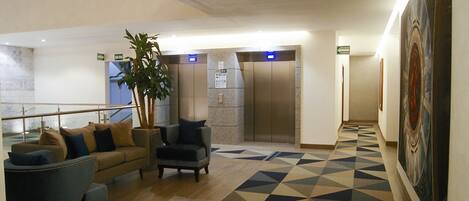 A lobby with a geometric patterned floor, a sofa, armchairs, a potted plant, and an elevator.