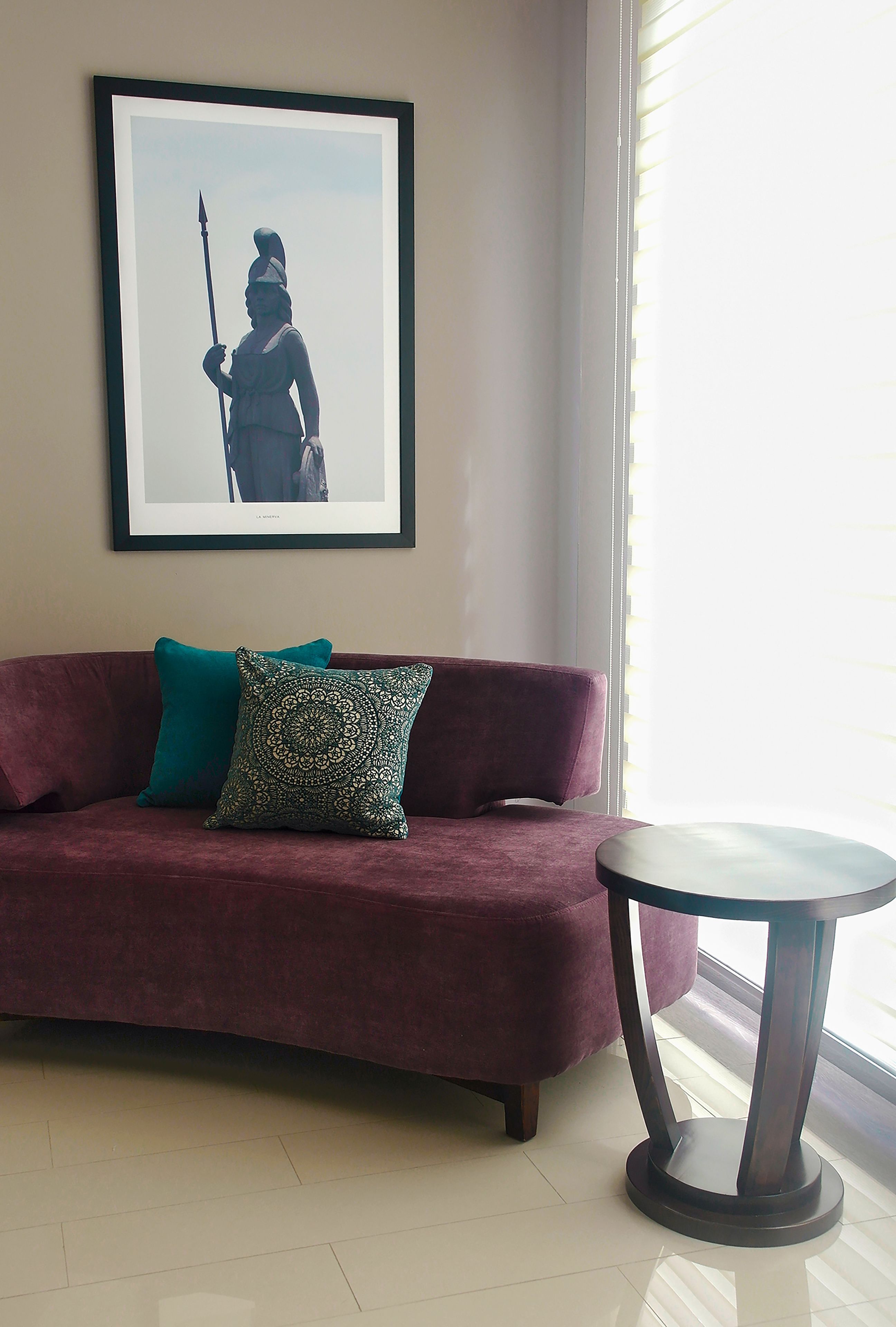 A room with a maroon sofa, two patterned cushions, a framed picture on the wall, and a round table.