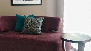 A room with a maroon sofa, two patterned cushions, a framed picture on the wall, and a round table.