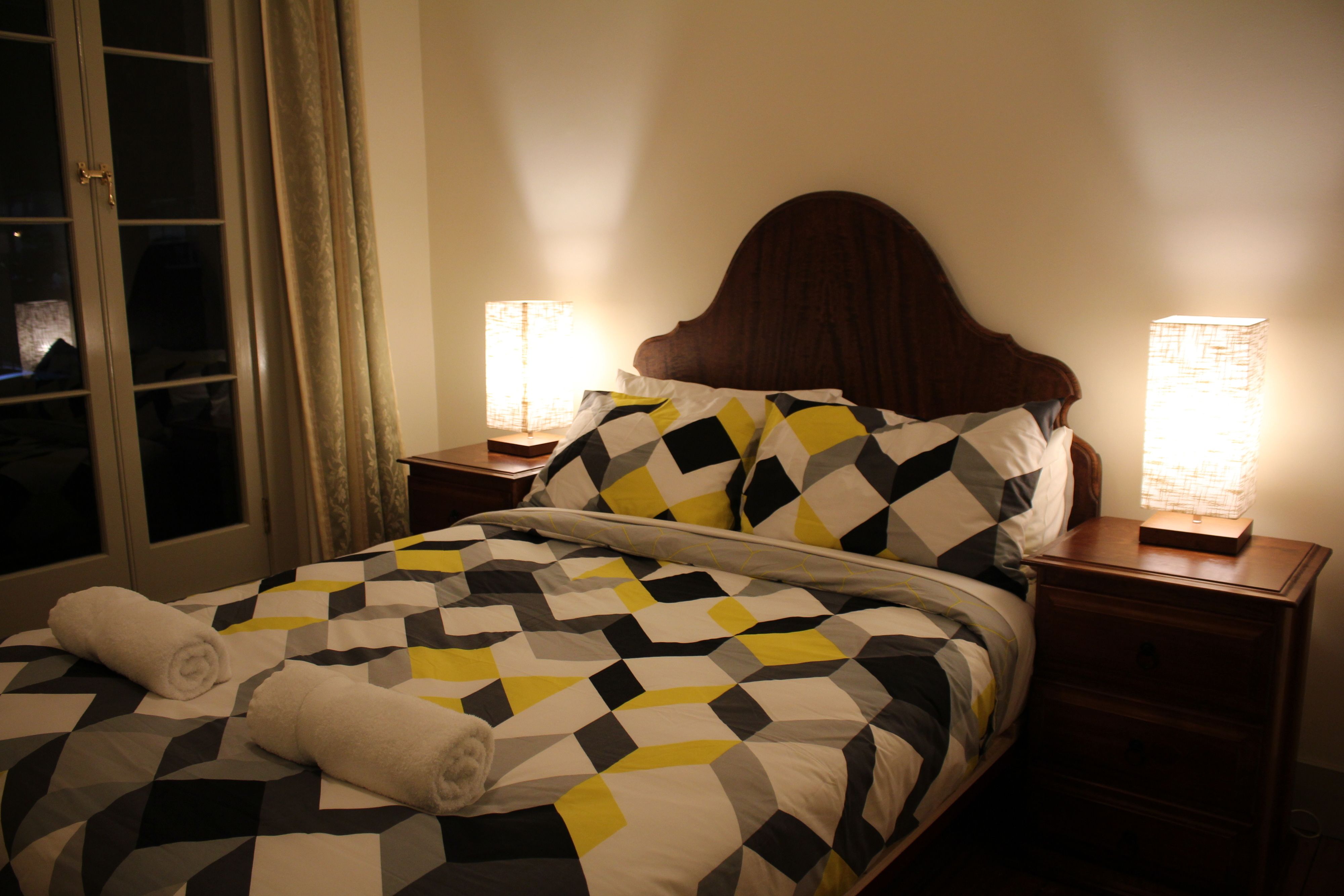 Cama bem arrumada com colcha estampada em padrão geométrico, duas luminárias de cabeceira e uma janela com cortinas.