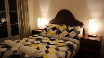 Cama bem arrumada com colcha estampada em padrão geométrico, duas luminárias de cabeceira e uma janela com cortinas.