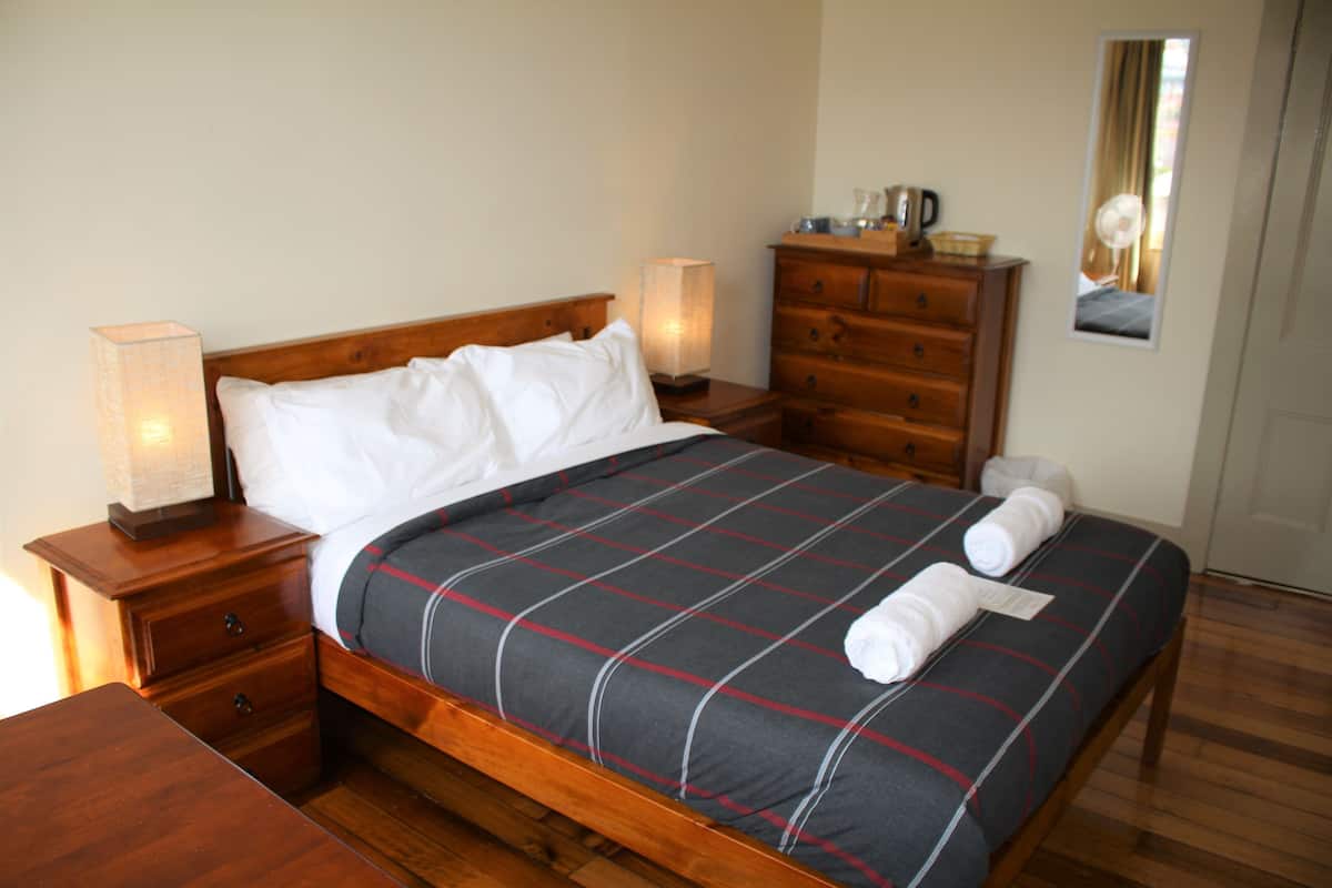 Siististi pedattu sänky, jossa on ruutukuosinen päiväpeite, puinen yöpöytä lampun kanssa, puinen lipasto ja peili seinällä.