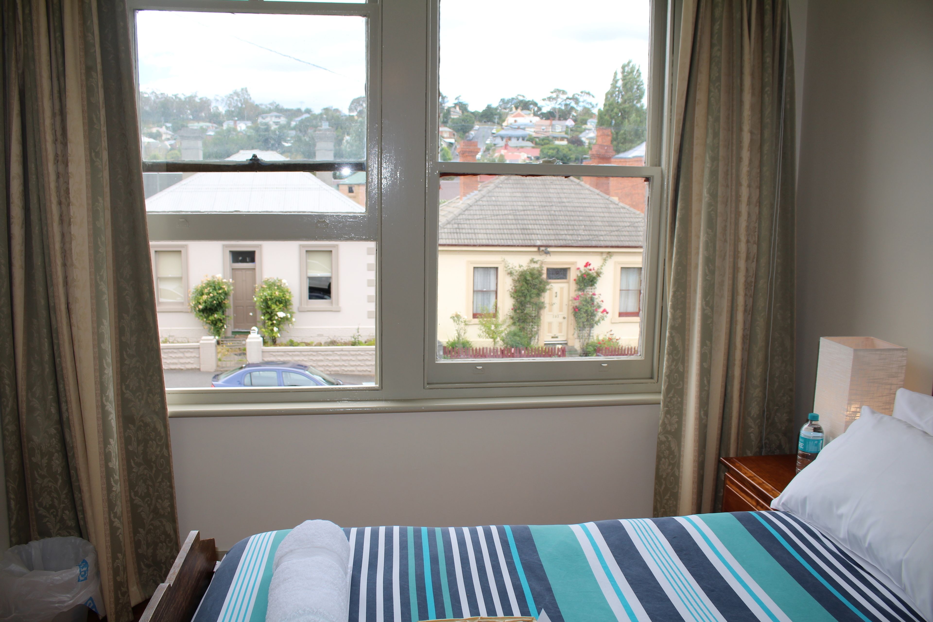 Une chambre d’hôtel avec un lit, une fenêtre avec des rideaux, une table de chevet avec une lampe, et une vue sur l’extérieur.