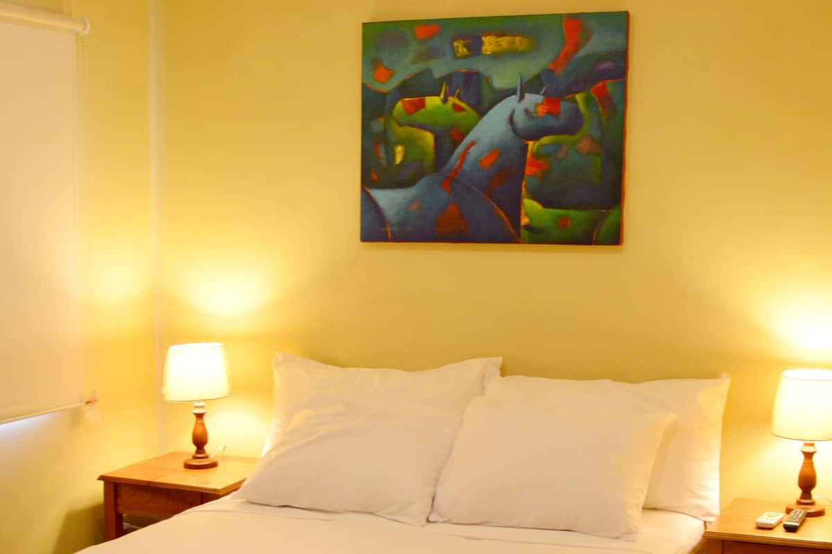 A hotel room with a bed, two bedside tables, a painting on the wall, and a green bedspread.