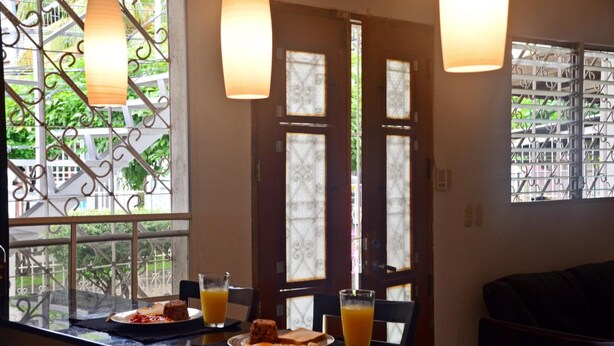 A dining area with a table set for breakfast, including plates with toast and orange juice, and a view of a garden through a window with decorative ironwork. - Hostal Santa Martha (Managua)