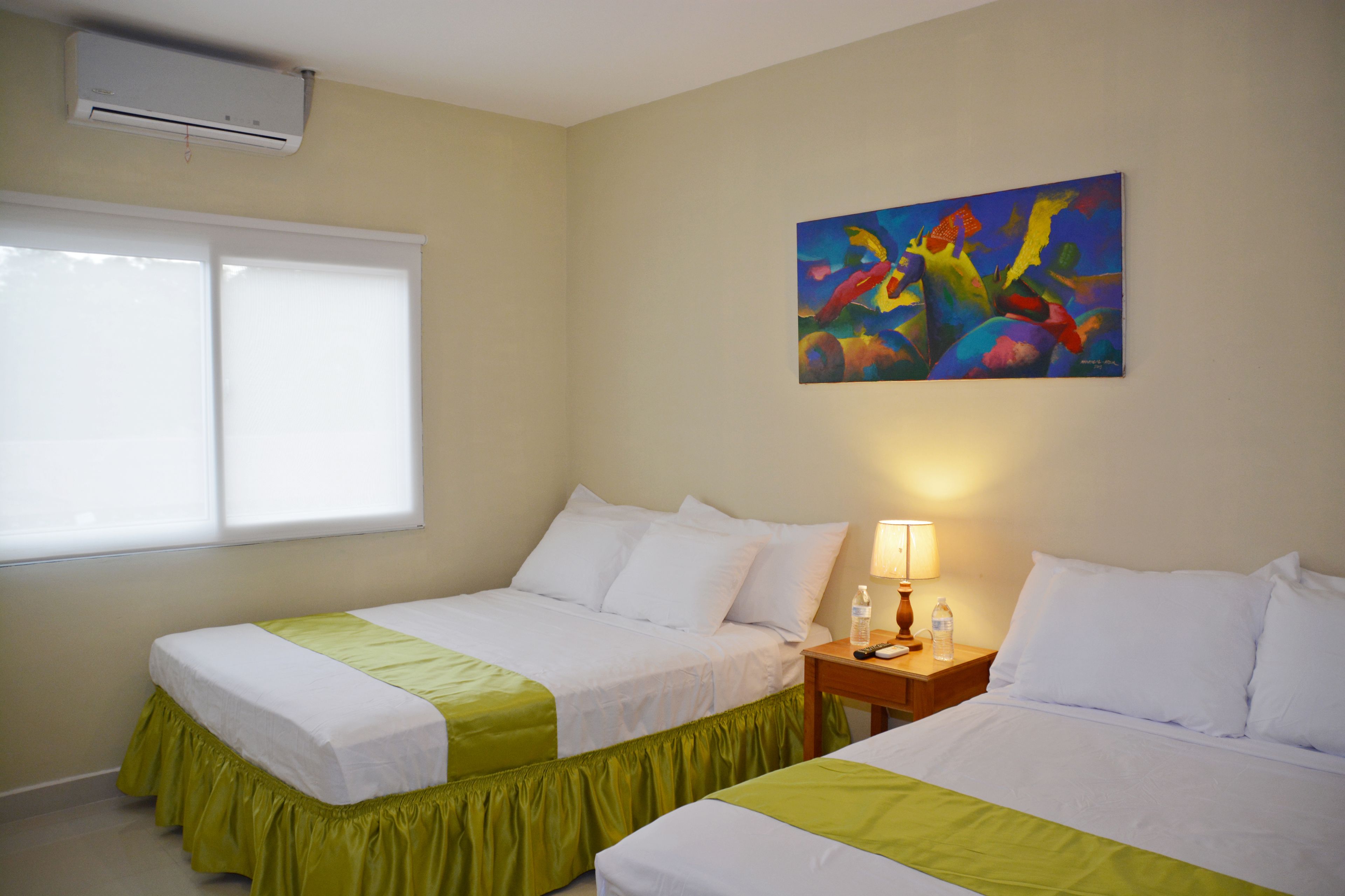 A hotel room with two beds, a window, an air conditioner, a painting, and a nightstand with a lamp and a bottle.