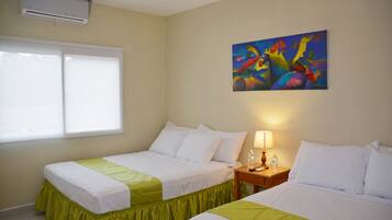 A hotel room with two beds, a window, an air conditioner, a painting, and a nightstand with a lamp and a bottle.