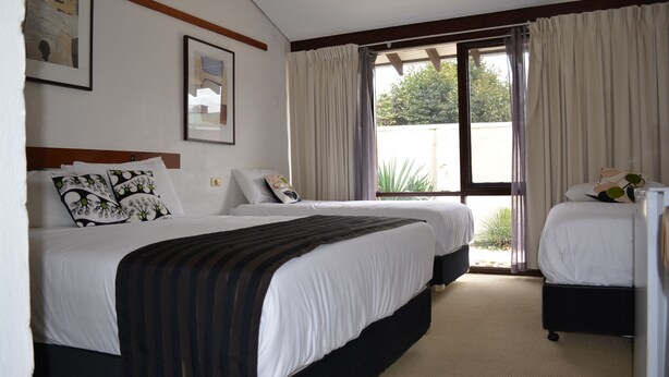 A hotel room with two beds, a large window with curtains, and a view of greenery outside. - The Apple Inn (Batlow)
