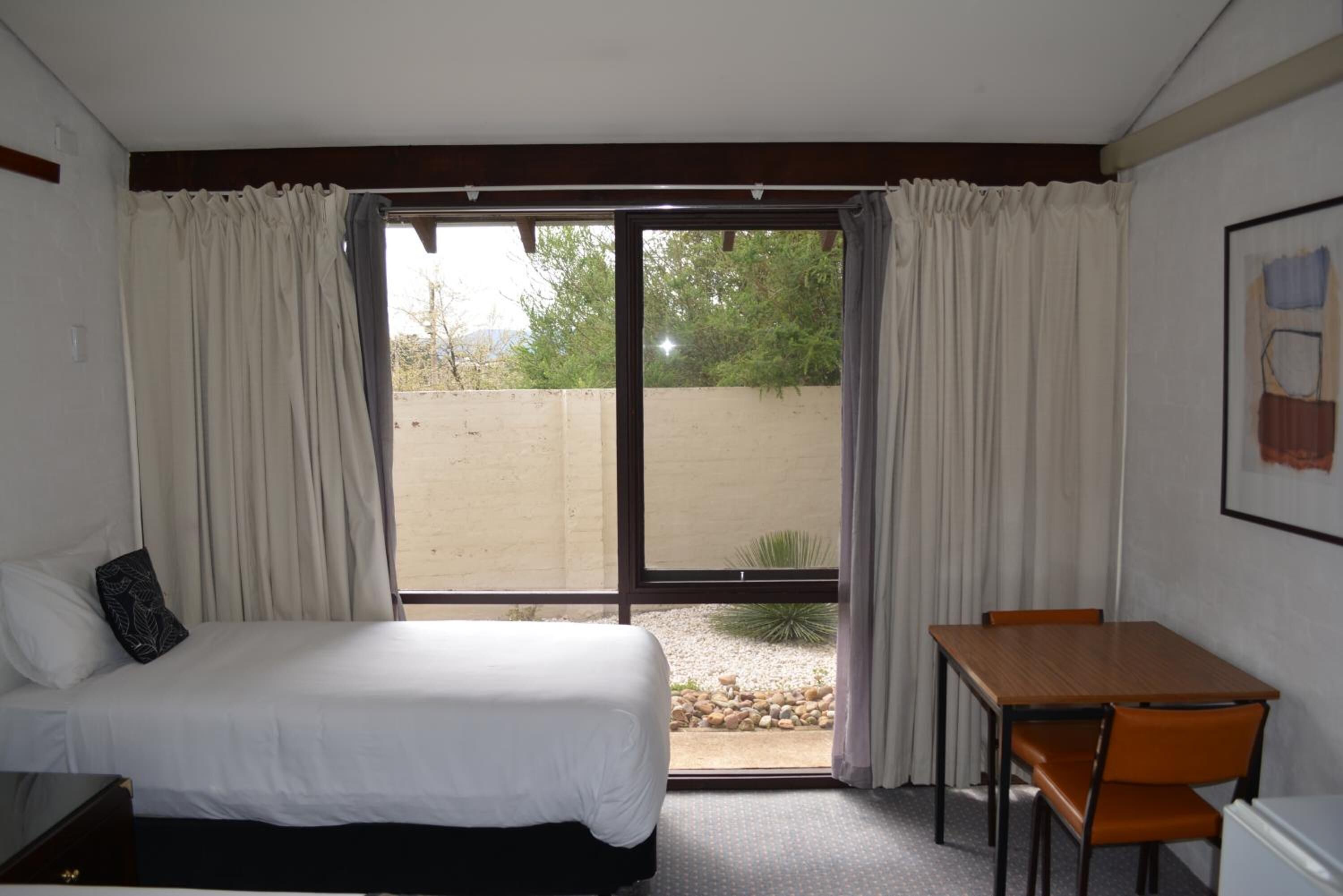 A hotel room with a bed, a table, and a chair. There is a sliding glass door leading to an outdoor area with a view of trees and a fence.
