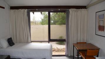 A hotel room with a bed, a table, and a chair. There is a sliding glass door leading to an outdoor area with a view of trees and a fence.