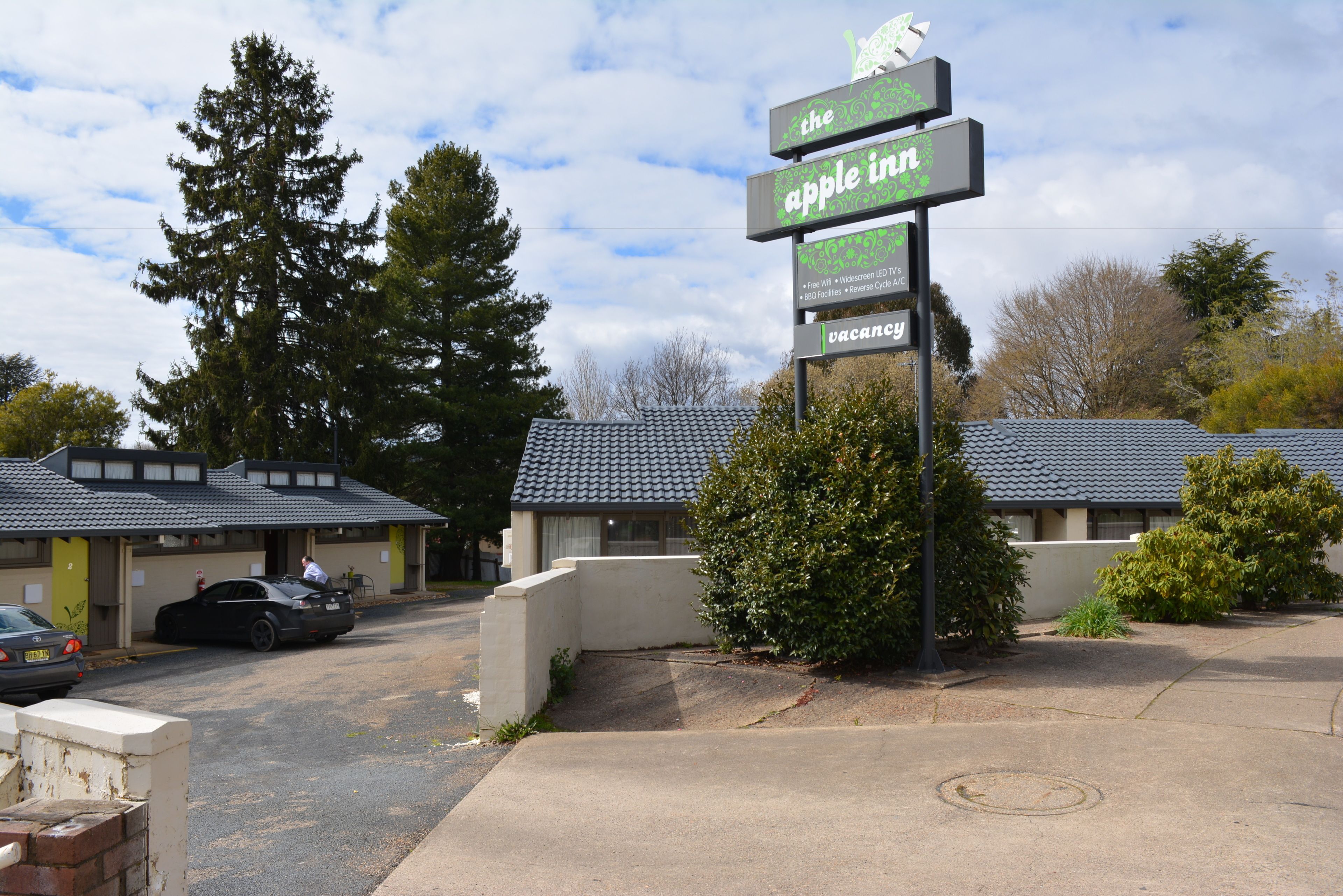 A motel with a sign indicating it is an Apple Inn, featuring a two-story building with a tiled roof, a parking area with a car, and a large tree in the background.