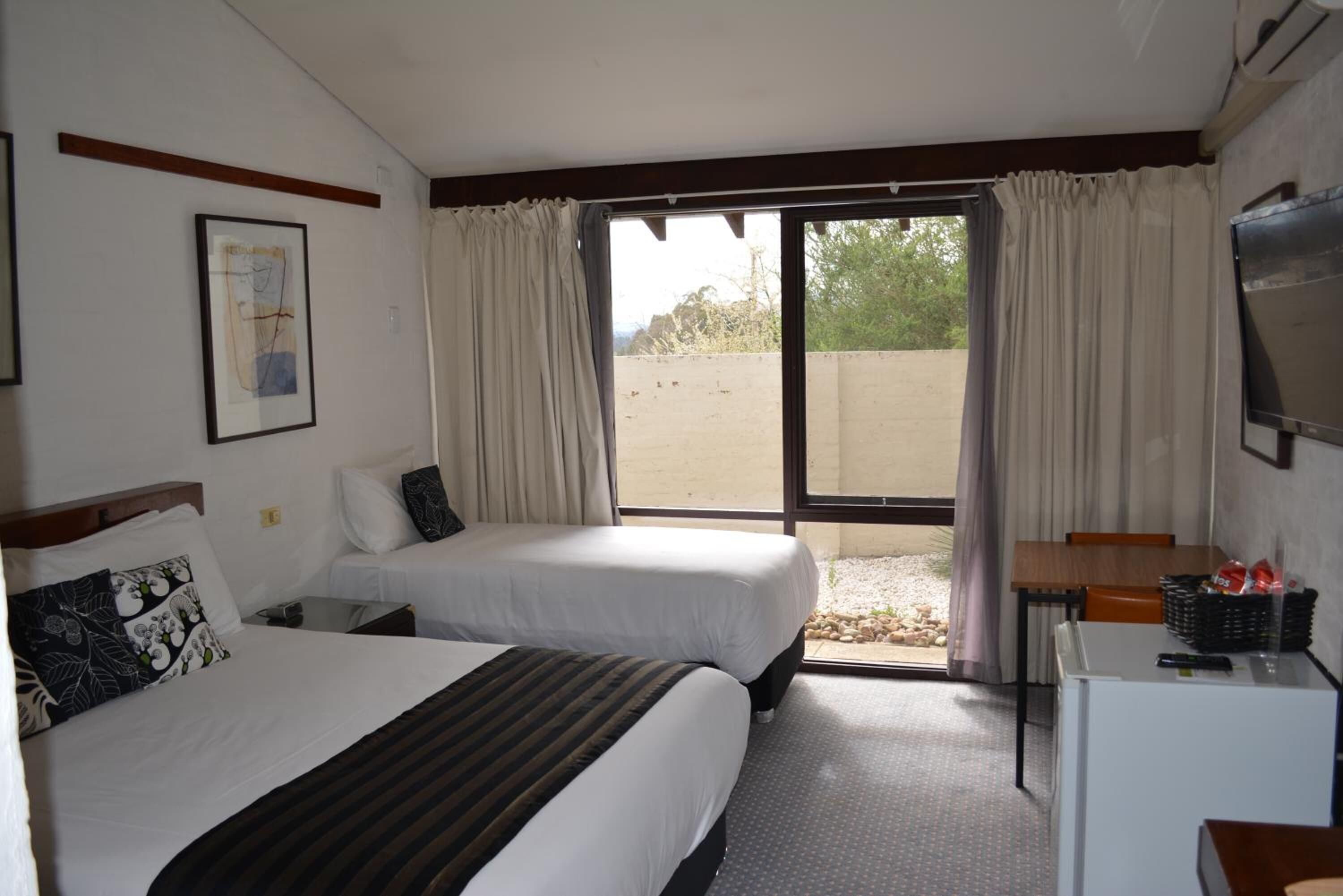 A hotel room with two beds, a small desk, a television, and a window with curtains.