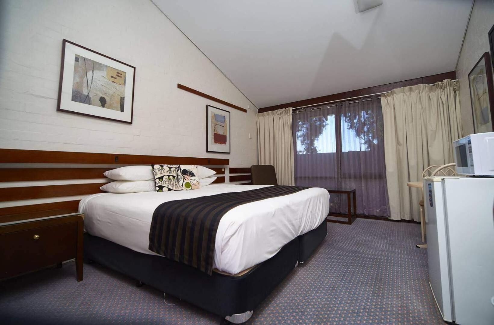 A hotel room with a bed, a desk, a chair, a microwave, and a refrigerator.
