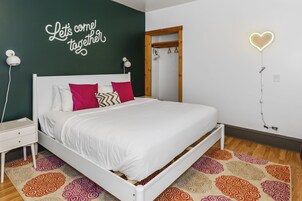 A neatly made bed with a white headboard, a wall with a 'Let's come together' sign, a heart-shaped light, and a wooden door frame.