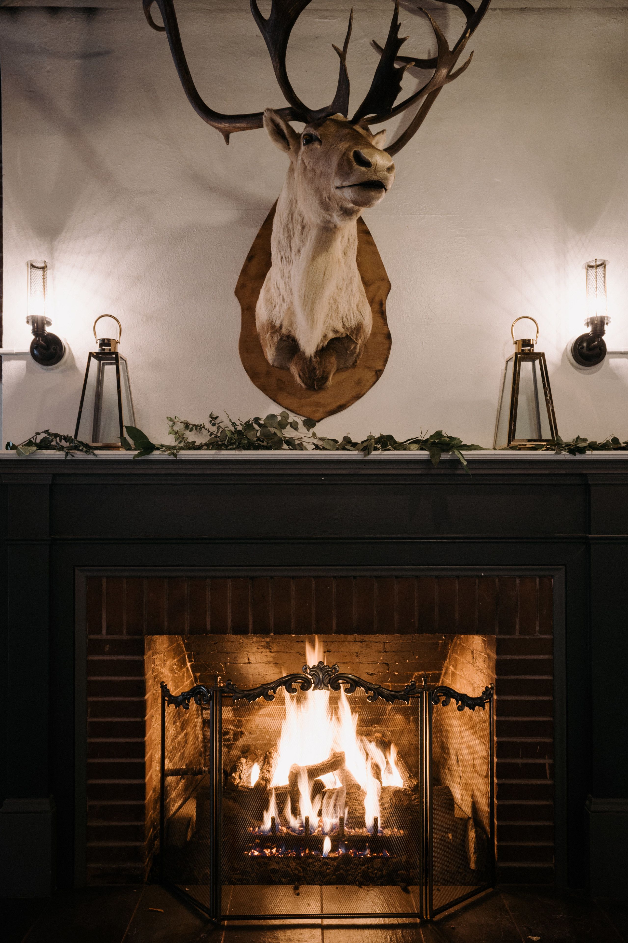 A fireplace with a mounted deer head above it, surrounded by greenery and two wall-mounted lanterns.