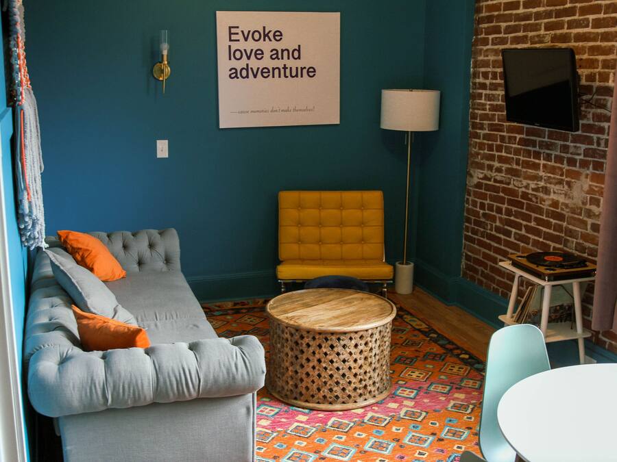 Una sala de estar acogedora con pared azul, un sillón amarillo, un sofá gris con cojines naranjas, una mesa de centro redonda de madera y una alfombra estampada.