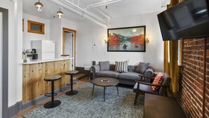 A modern living room with a sofa, coffee table, and bar stools. There is a flat-screen TV mounted on the wall, a painting above the sofa, and a kitchen area with a refrigerator and microwave. - Hood River Hotel (Hood River)