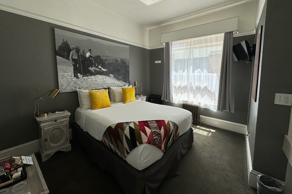 A hotel room with a bed, a nightstand, a lamp, a painting, a window with curtains, and a television.