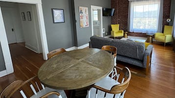 A dining area with a round wooden table and white chairs, a gray sofa, and a brick wall with a TV.