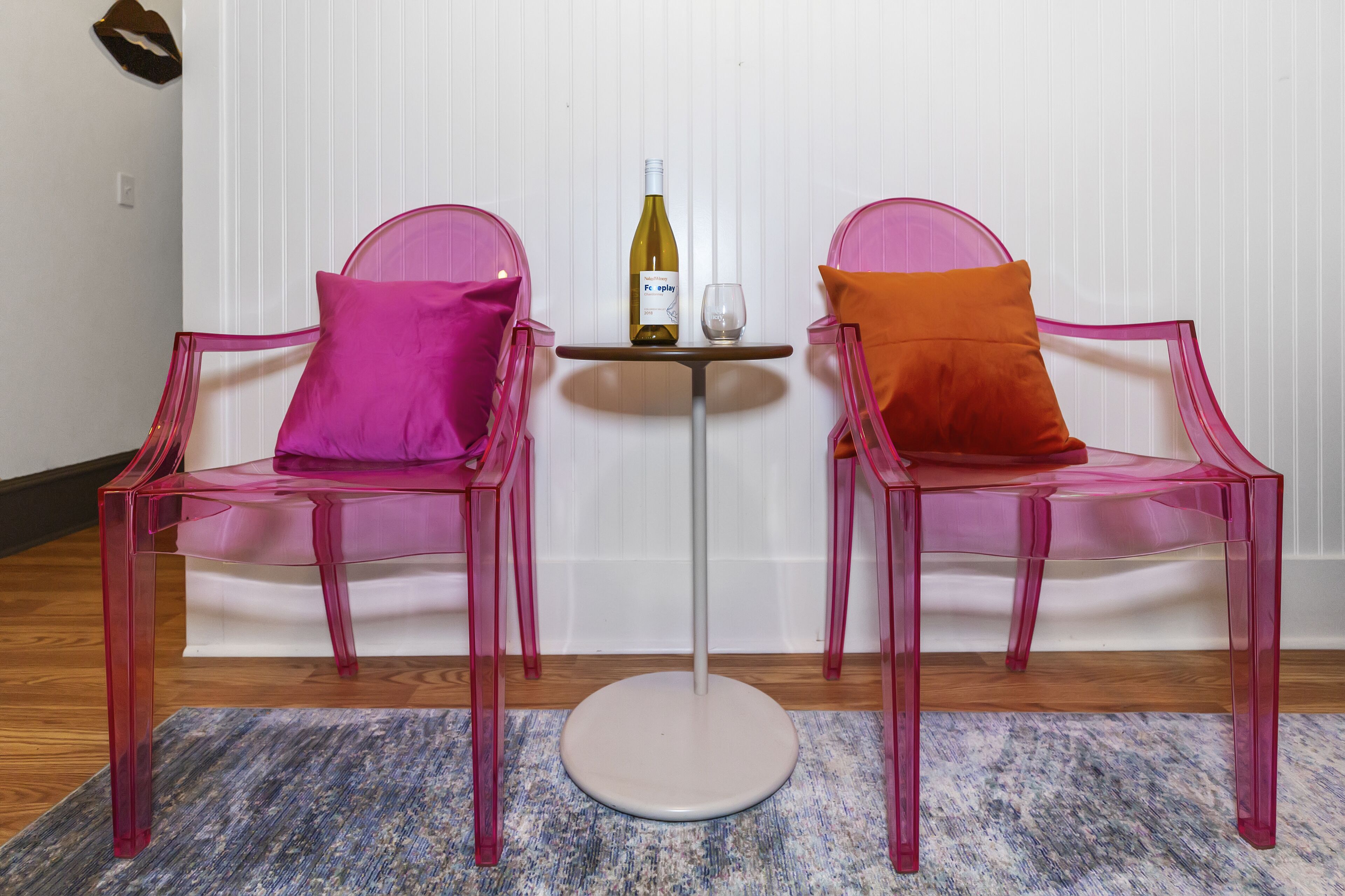 Two transparent plastic chairs with cushions, a small round table with a bottle and glass, and a patterned rug on a wooden floor.