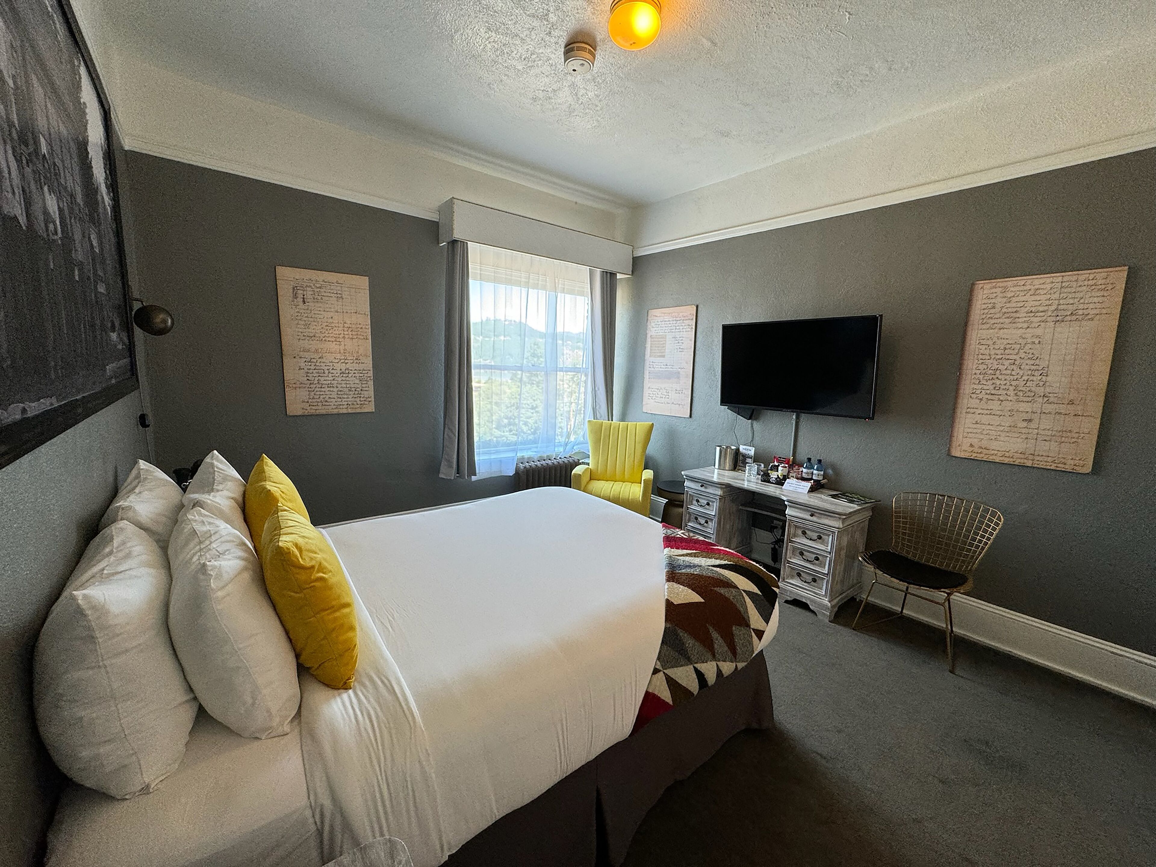A hotel room with a large bed, a desk, a chair, a TV, and two windows.