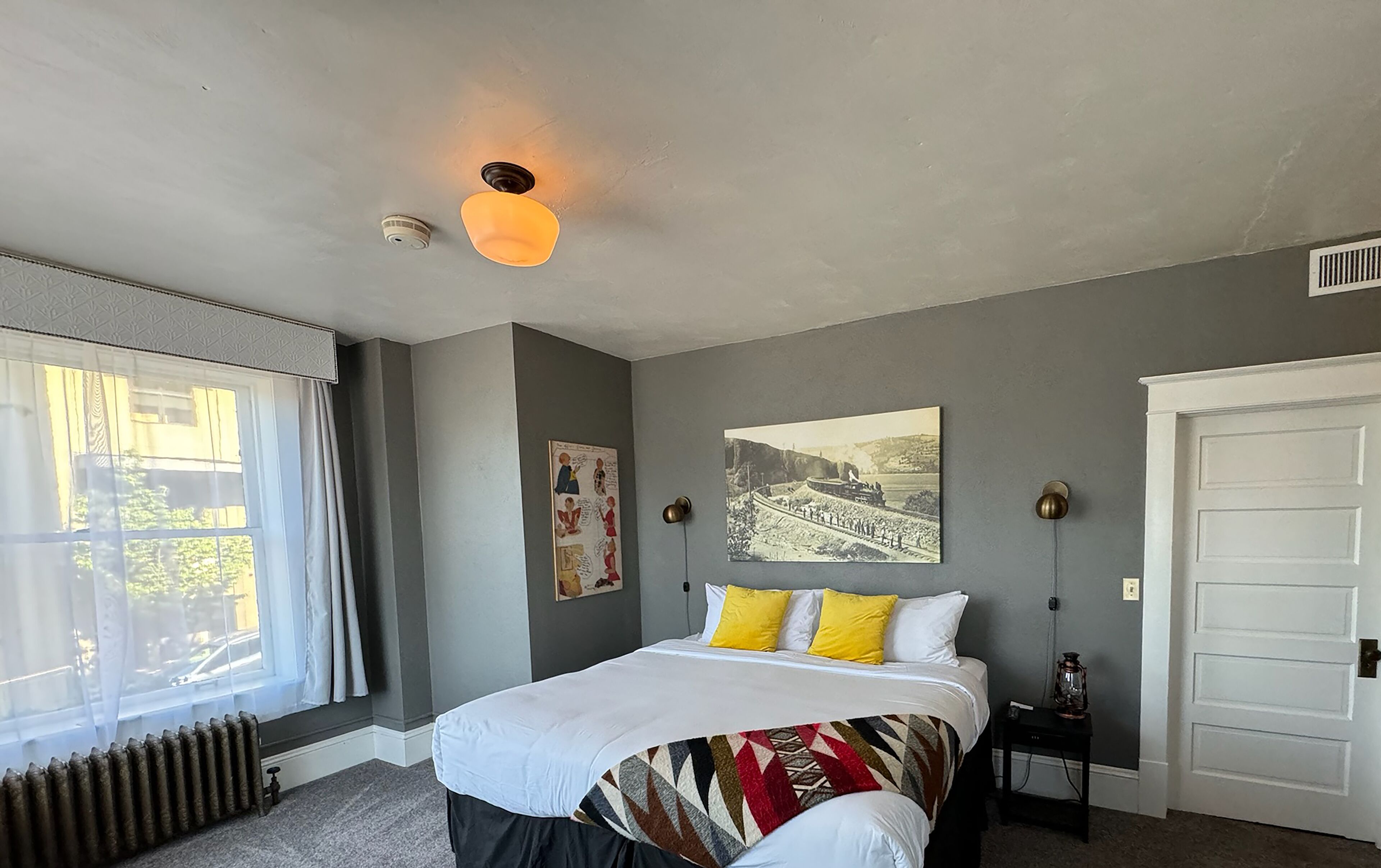 A bedroom with a bed, a radiator, a window with curtains, a door, and a wall with a picture.
