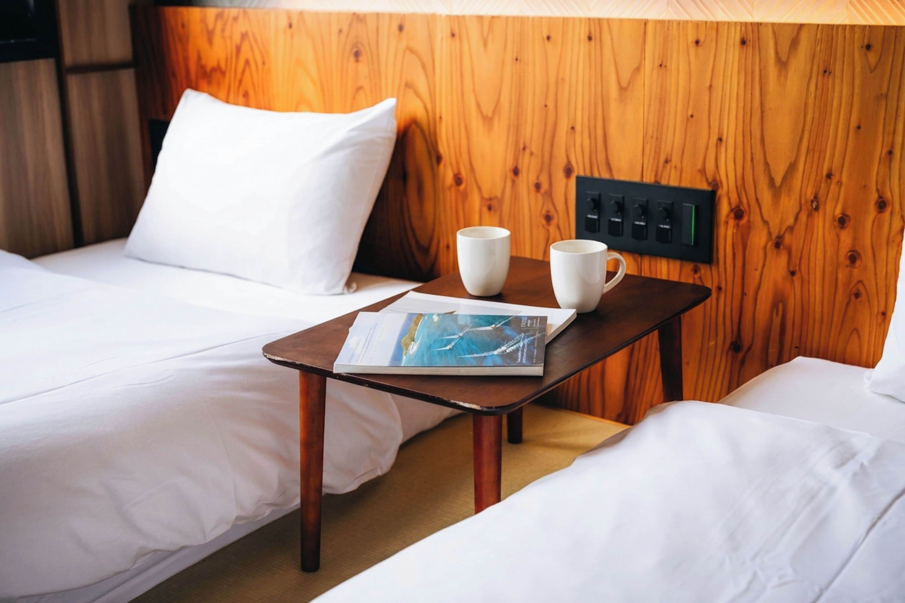 Habitación de hotel con dos camas, cabecero de madera, una mesita con una revista y dos tazas, y un panel de control en la pared.