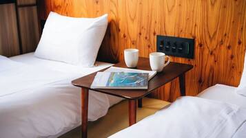 Habitación de hotel con dos camas, cabecero de madera, una mesita con una revista y dos tazas, y un panel de control en la pared.