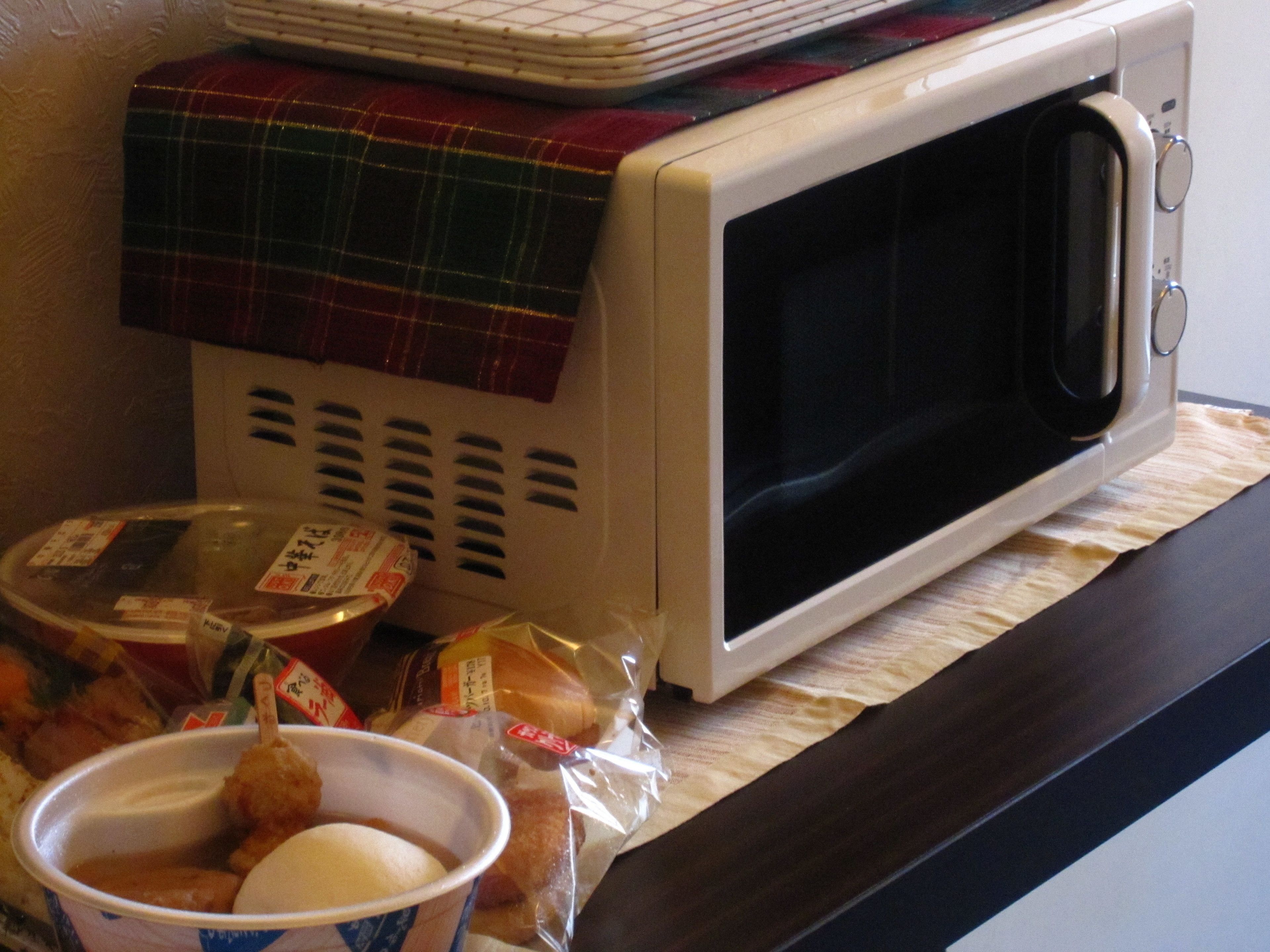 A microwave oven on a kitchen counter with a plaid cloth over it, a bowl of food, and some packaged food items.