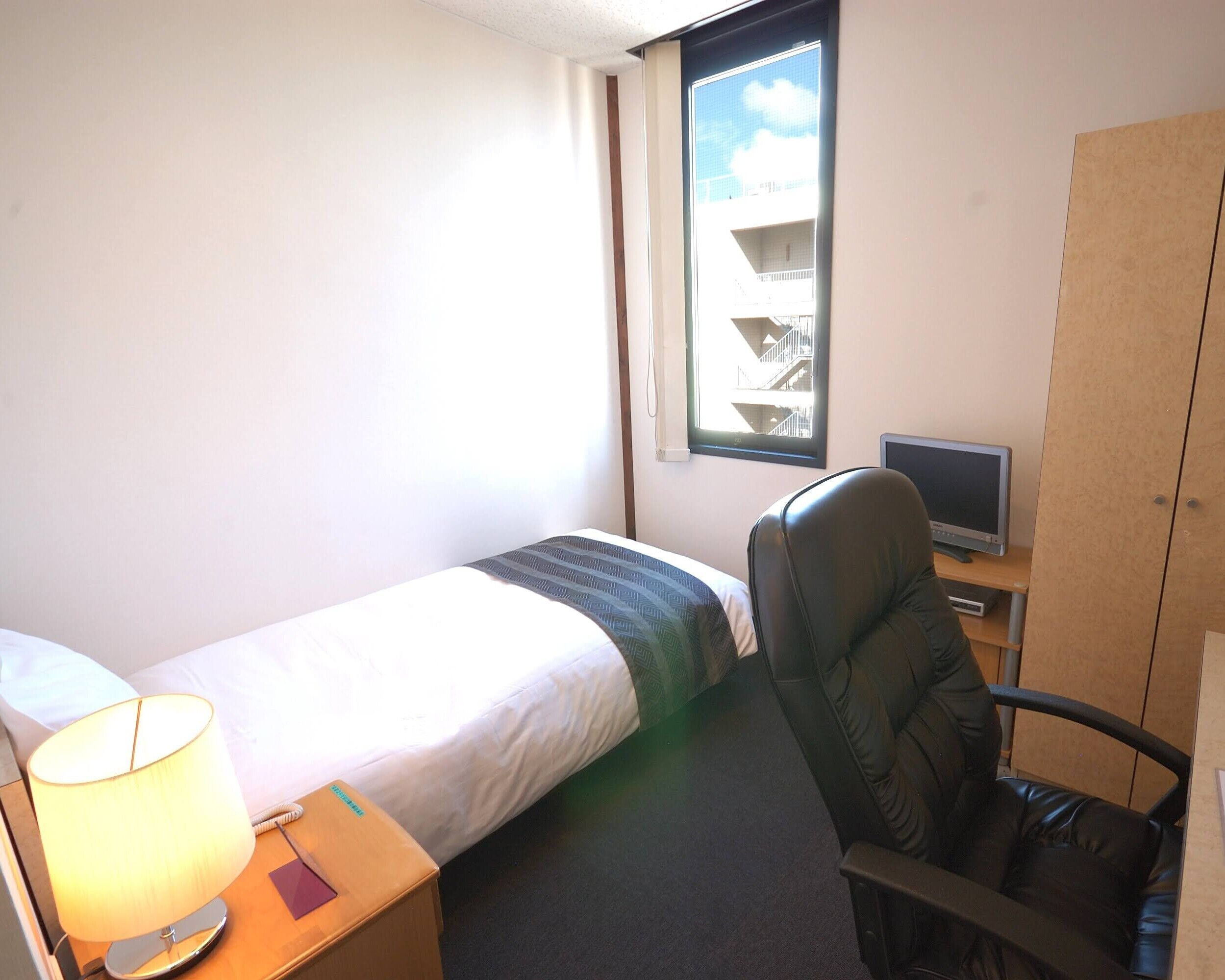 A hotel room with a bed, a desk with a lamp, a chair, a TV, and a wardrobe.